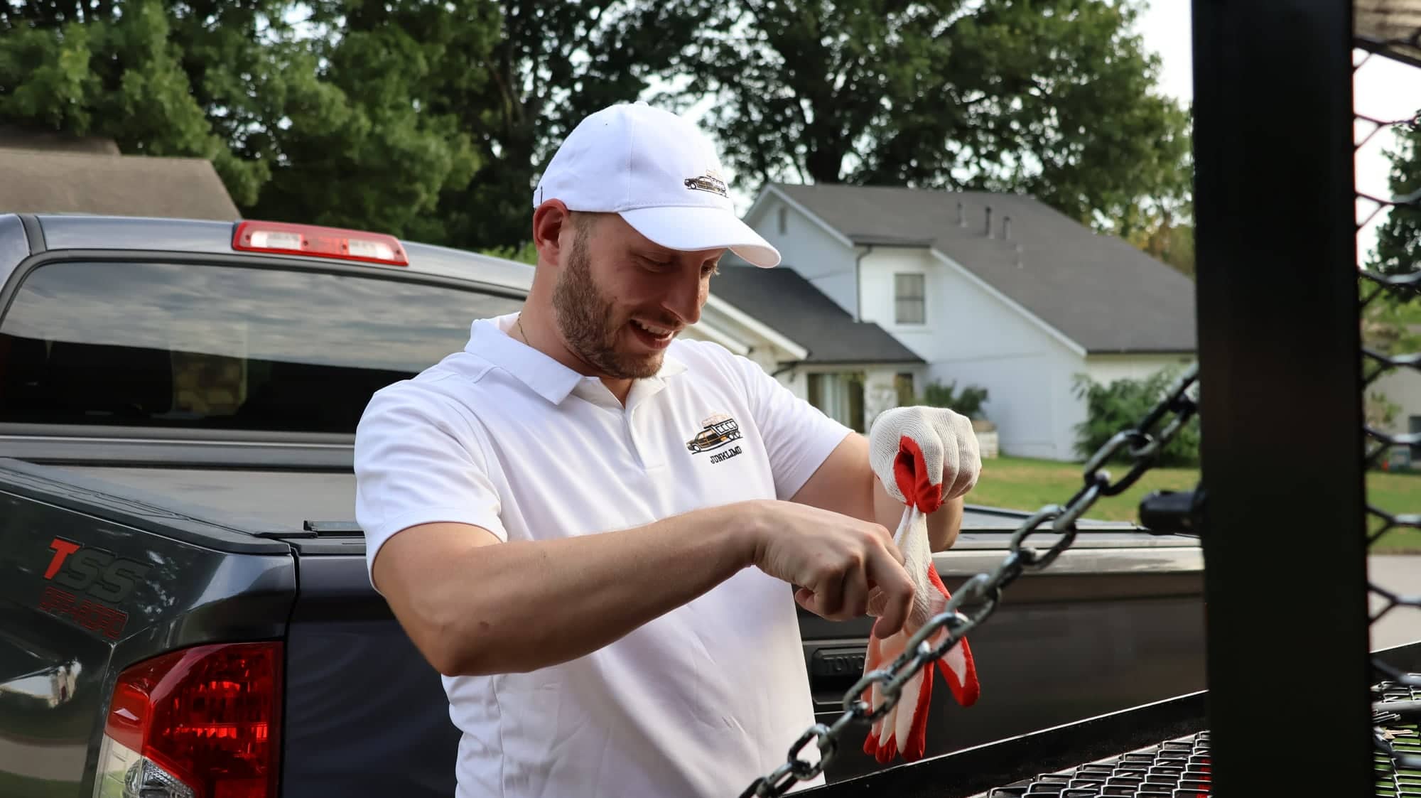 Victor pulling on red and white work gloves at the open tailgate of his pickup truck outside a Tulsa house, wearing a white polo and cap with the JunkLimo logo